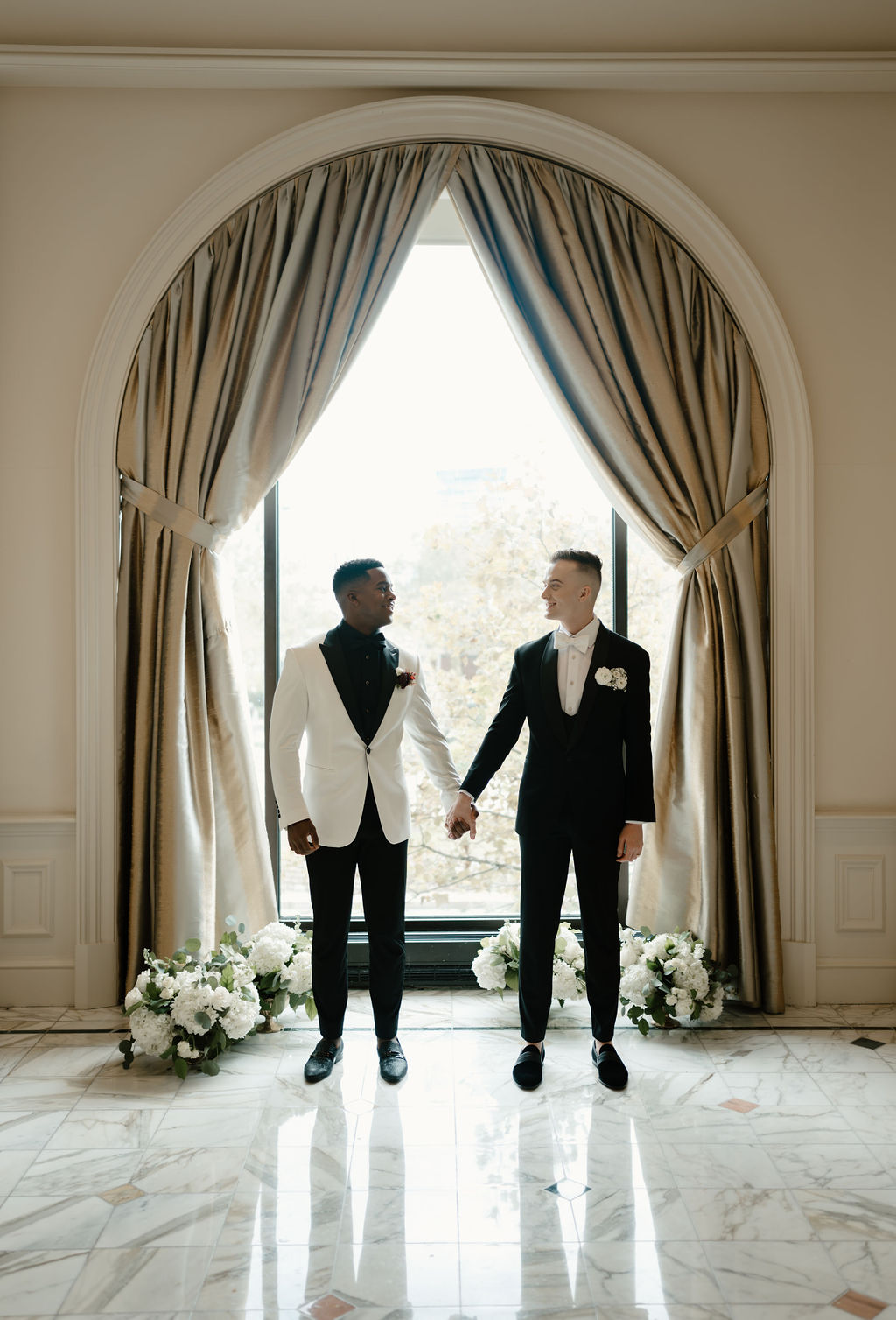 Two men holding hands and looking into eachothers eyes in front of arched window,